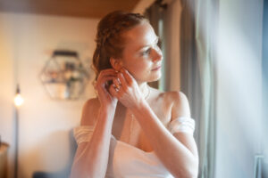 The bride looks out the window while putting on her earrings