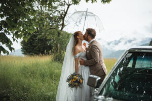 Bride and groom share a kiss beside the Cadillac beneath an umbrella