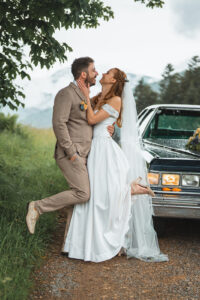 The couple stands in front of a Cadillac, lifting their legs playfully while laughing together