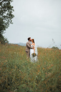 The groom kisses his bride on the forehead in a field of golden grass beneath a cloudy sky