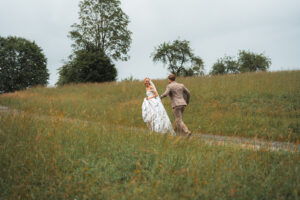 The couple walks uphill through tall grass and orange flowers, smiling at each other