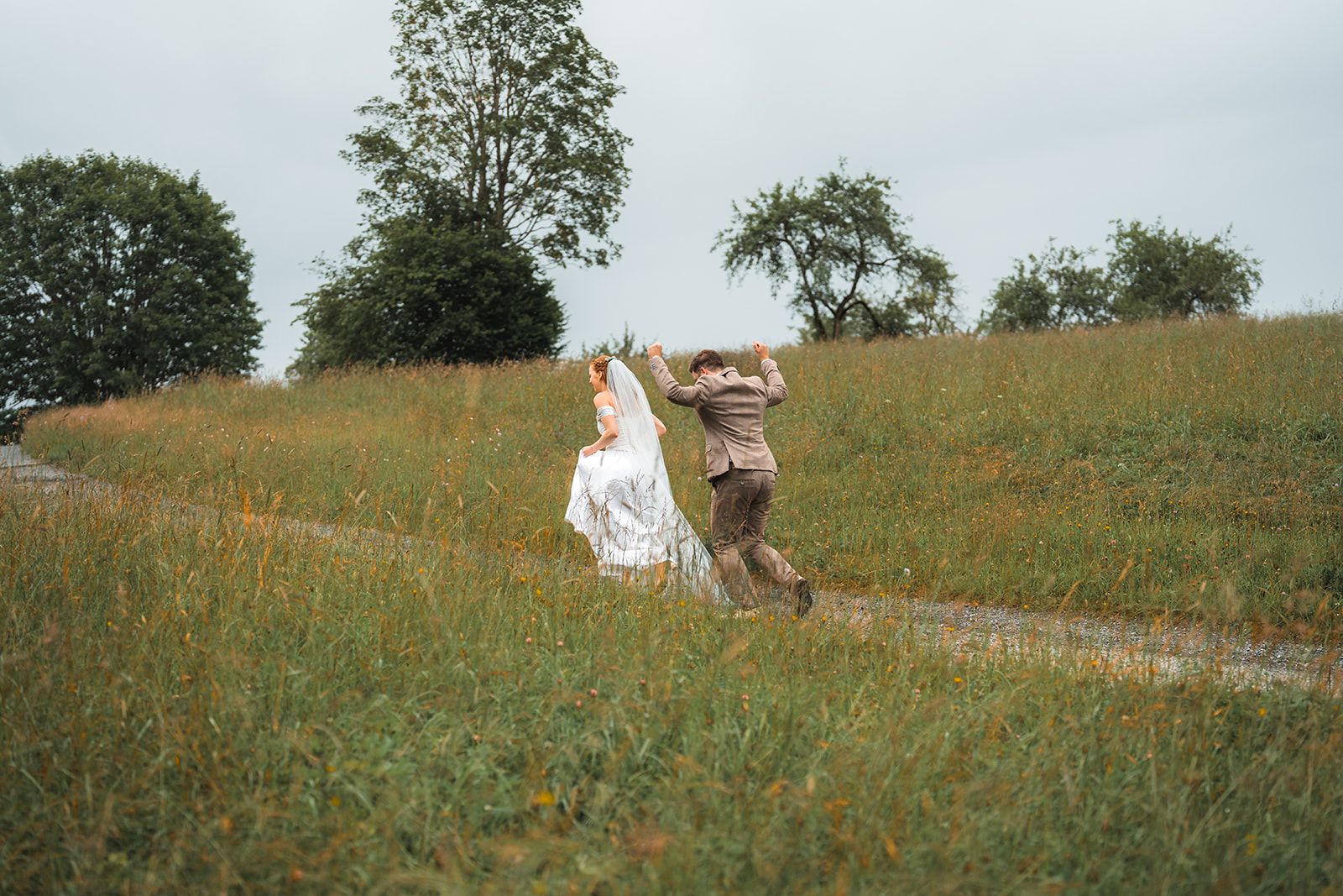 Wedding couple joyfully running up a path through tall green grass and orange flowers