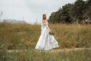 Full-body portrait of the bride standing in a field, looking down at her dress