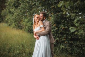 The groom hugs his bride from behind and kisses her cheek in front of a hedge
