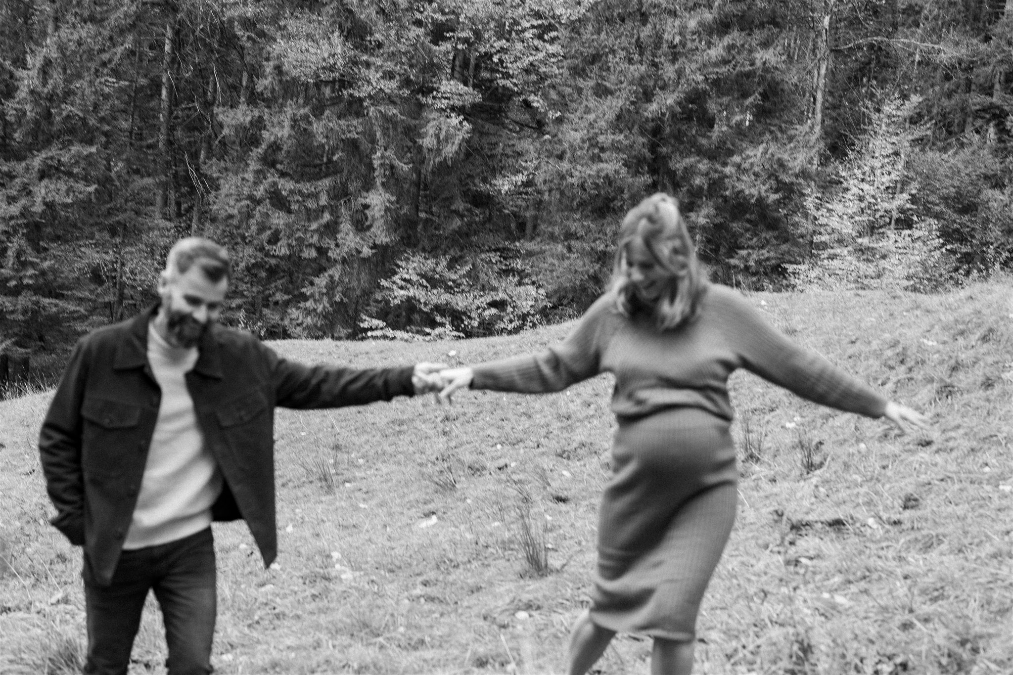 Black-and-white photo of an expecting couple walking hand in hand through a meadow