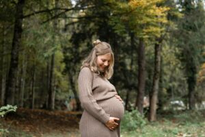 Schwangere Frau steht vor Herbstbäumen, hält ihren Babybauch und blickt darauf