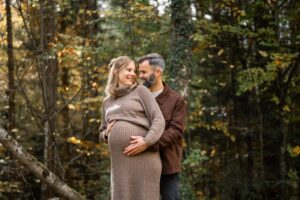 Schwangeres Paar steht vor Herbstbäumen und hält gemeinsam den Babybauch und lacht