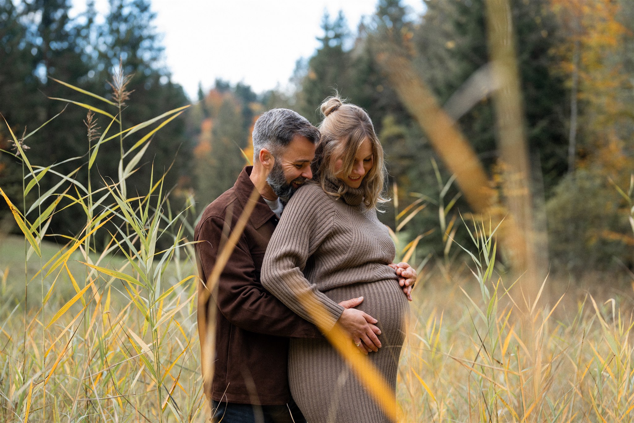 Pregnant couple standing in tall grass while the man embraces the woman from behind and holds her baby bump