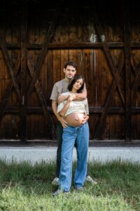 Man embracing his pregnant wife from behind in front of a barn while both look at the camera