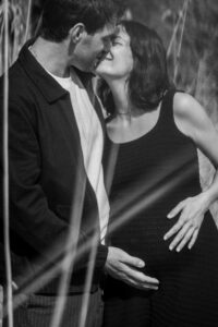 Black-and-white photo of a couple kissing in tall grass with hands resting on the baby bump
