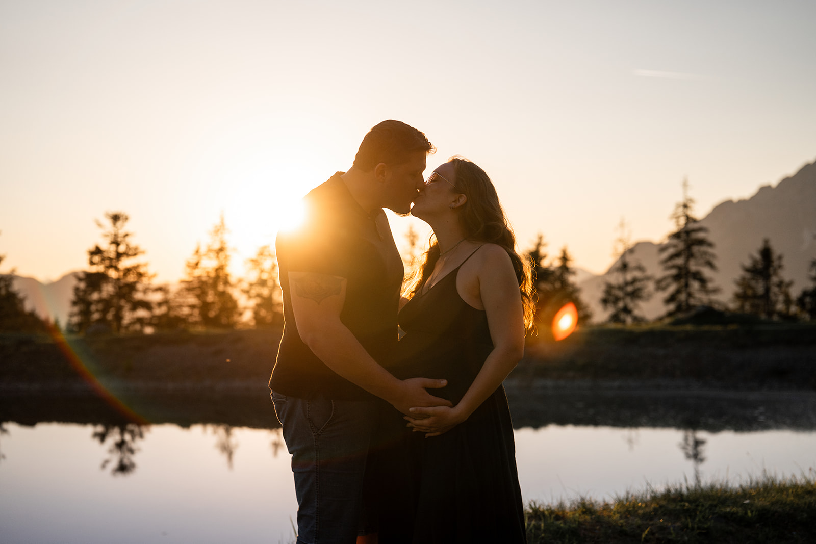 Pregnant couple kissing at sunset by a mountain lake while holding their hands on the baby bump