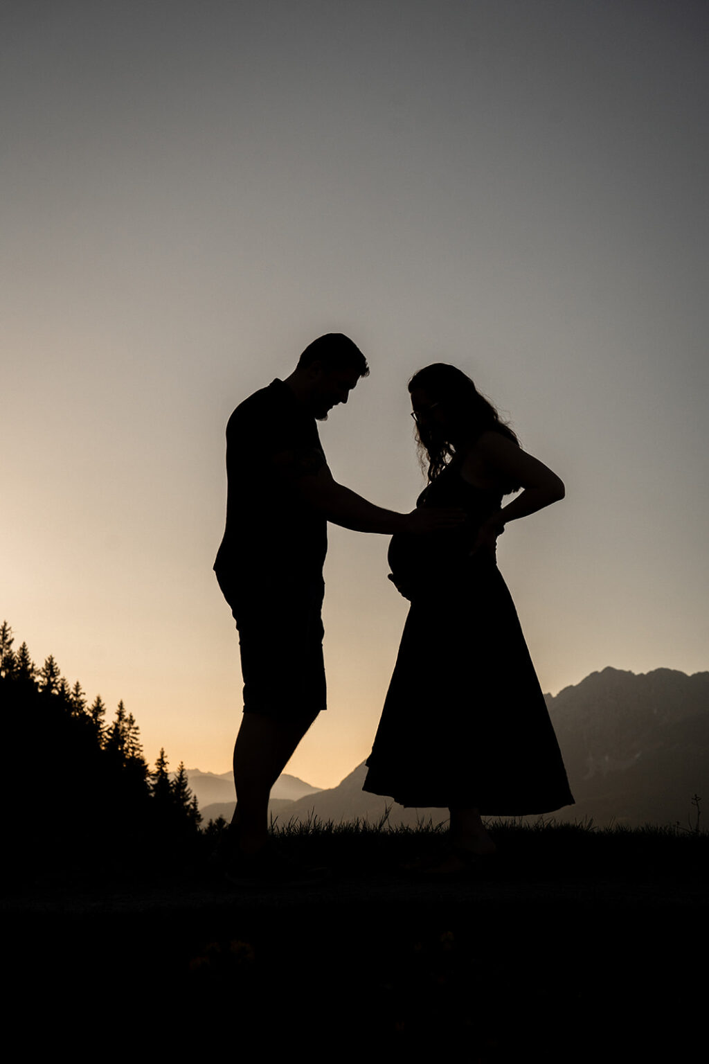 Silhouettenfoto eines schwangeren Paares bei Sonnenuntergang, der Mann legt seine Hand auf den Babybauch