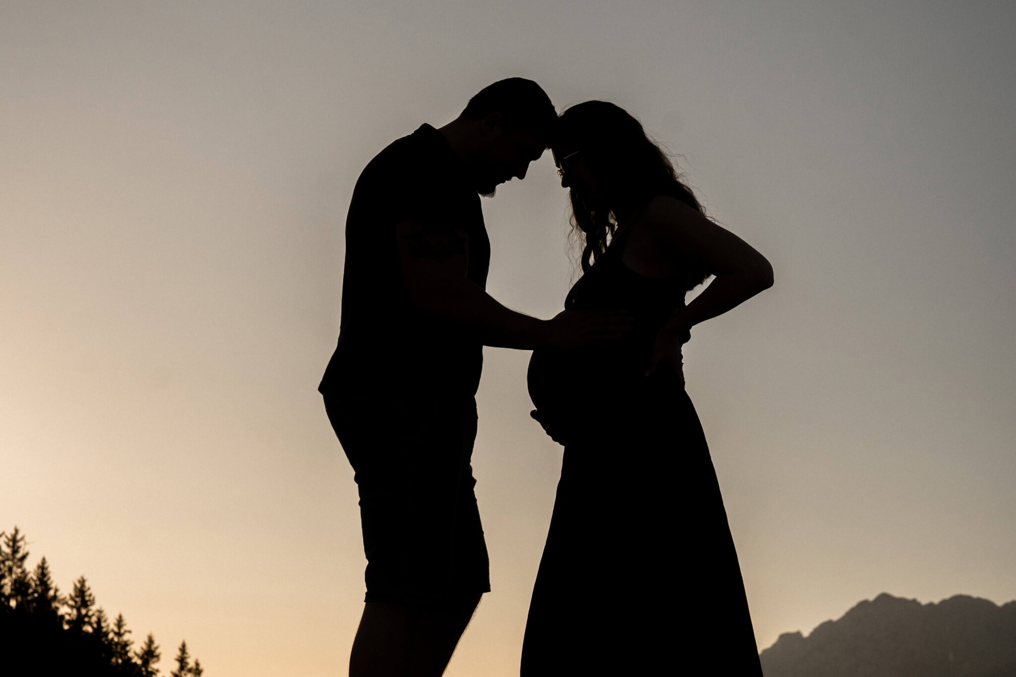 Silhouette photo of a pregnant couple at sunset while the man places his hand on the baby bump