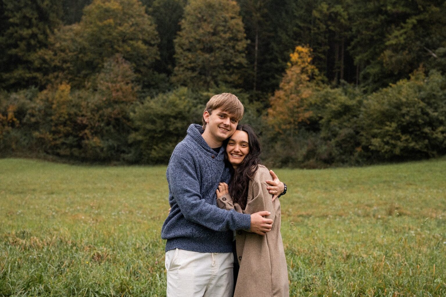Young couple embracing in a field and smiling at the camera with an autumn landscape behind them