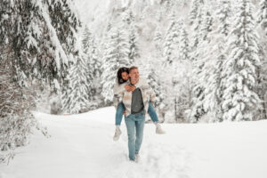 Austrian-Thai couple walking piggyback through a snowy field while the woman smiles playfully over his shoulder