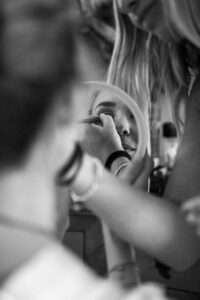Black-and-white close-up of the bride applying makeup in a handheld mirror