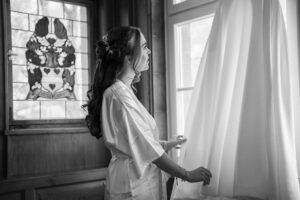 Black-and-white photo of the bride looking at and touching her wedding dress by the window