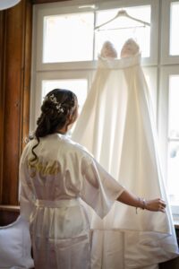 The bride stands by the window in a “Bride” robe holding her wedding dress