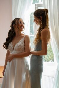 Bride and maid of honor standing by the window, smiling at each other