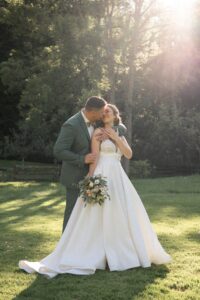 The wedding couple stands in golden light as the groom hugs his bride from behind and kisses her cheek