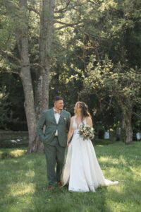 The wedding couple walks hand in hand across a meadow beneath a tree
