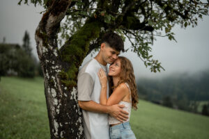 Engaged couple standing beneath a tree while the woman looks up at her partner