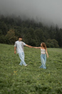 Young woman leading her fiancé across a meadow while both smile at each other