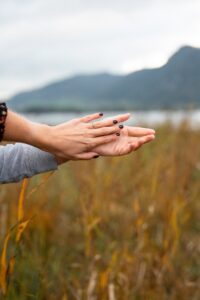Close-up of hands holding each other in front of orange grass, a lake, and mountains with a visible engagement ring