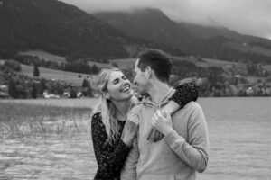 Black-and-white photo of a woman embracing her fiancé from behind by a lake while smiling warmly at him