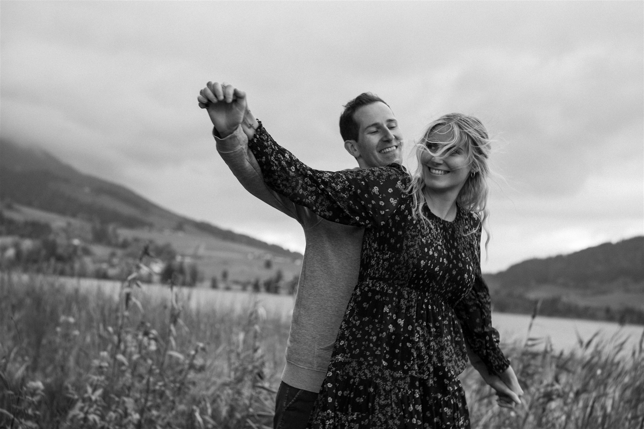 Black-and-white photo of an engaged couple in tall grass by a lake, while laughing and playing with their arms in the wind