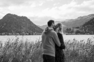 Black-and-white photo of a young couple in front of a lake and mountains with focus on the landscape