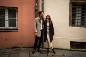 Couple leaning against each other in front of an old town building while wearing trench coats and sunglasses