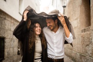 Young couple laughing beneath a trench coat in the old town while sheltering from the rain