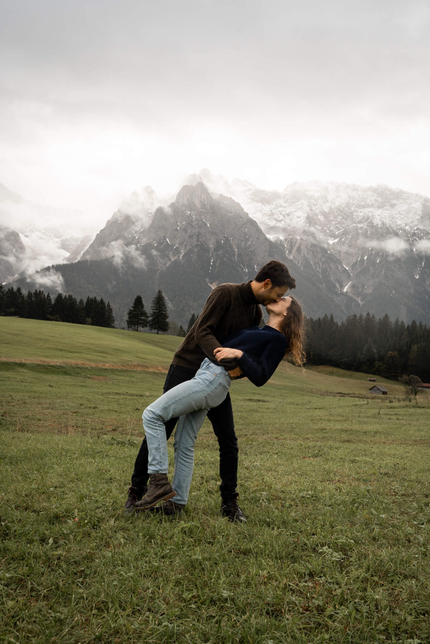 Man dipping his fiancée backward for a kiss in a green meadow with snowy mountains in the background