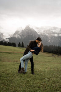 Man dipping his fiancée backward for a kiss in a green meadow with snowy mountains in the background