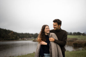 Man wrapping his jacket around his girlfriend while both laugh in front of a lake