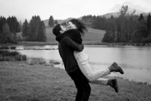 Black-and-white motion blur photo of a man lifting his fiancée in front of a lake and trees