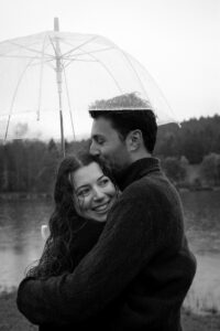 Medium close-up black-and-white photo of a couple under an umbrella while the man kisses his fiancée on the head
