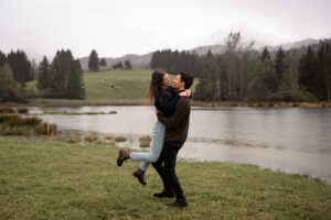 Full-body photo of a man lifting and twirling his fiancée in front of a lake and trees