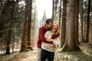 Man embracing his fiancé from behind while both laugh warmly in the forest