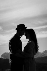 Black-and-white silhouette photo of a couple with their foreheads touching in front of mountain silhouettes and wispy clouds