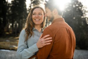 Engaged couple standing in golden light beneath trees while the woman laughs and rests her hand on his shoulder
