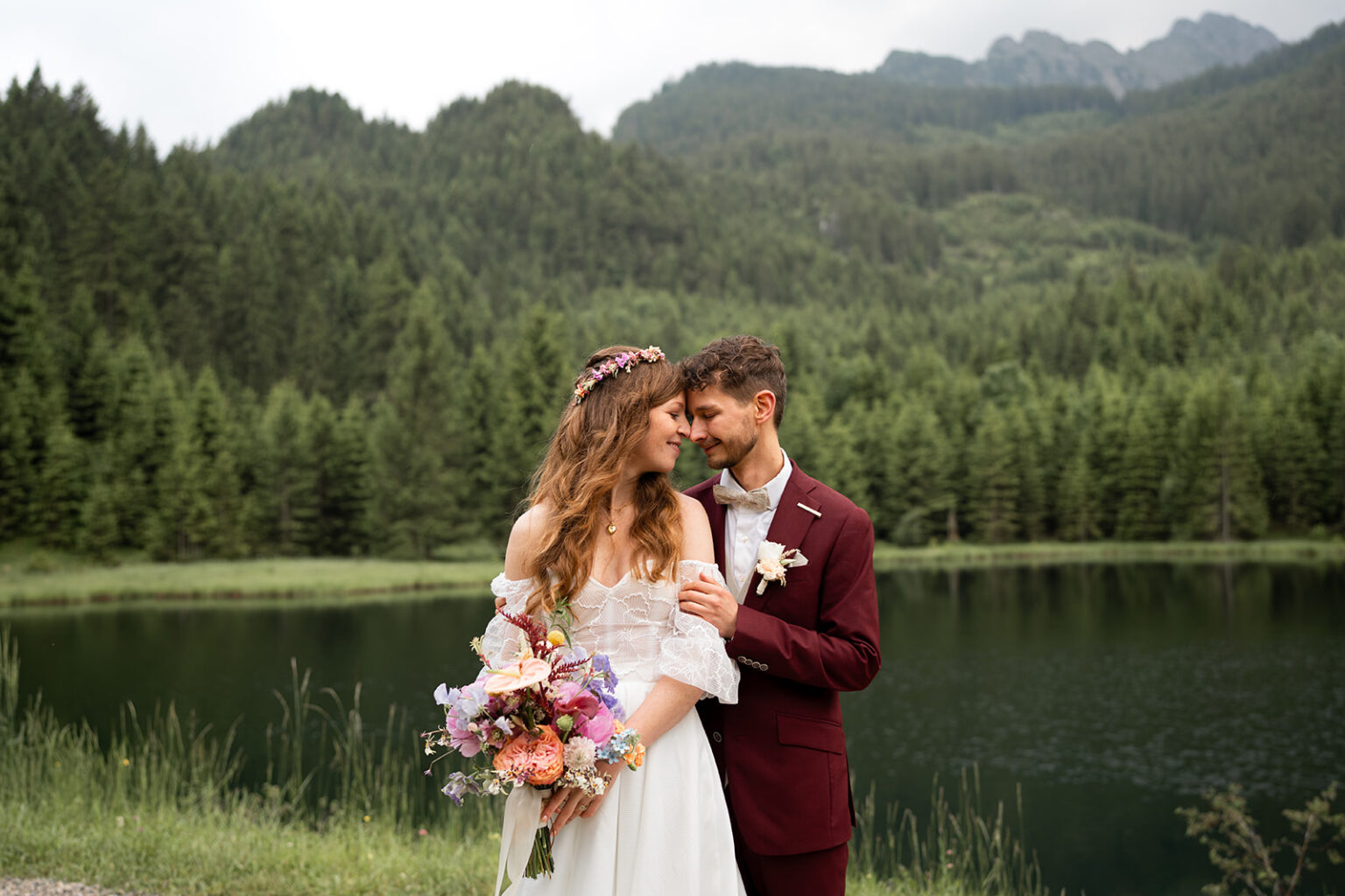 Young wedding couple by a lake with mountains in the background, leaning their heads together and smiling