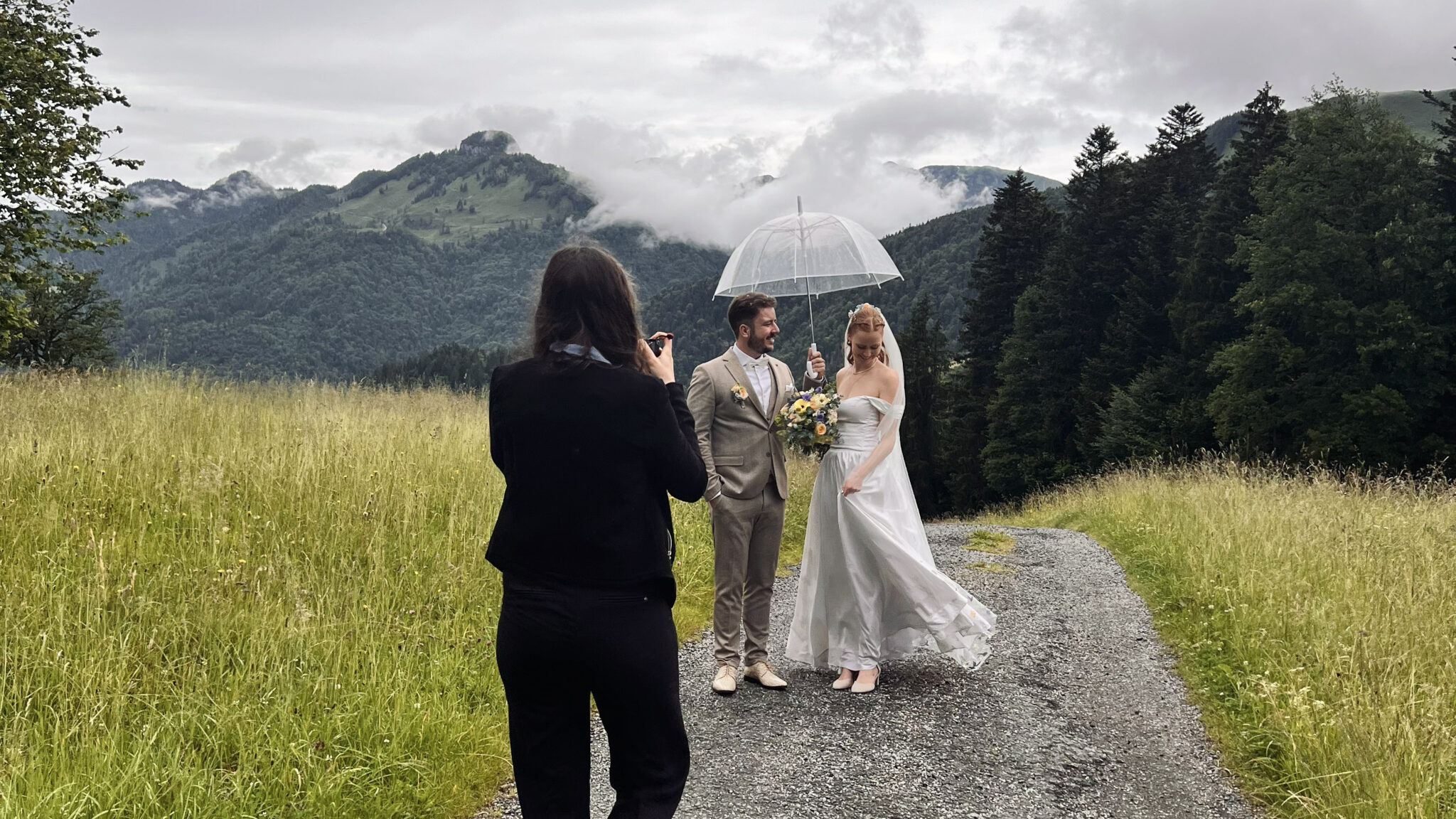 Photographer Caro photographing a happy married couple walking under an umbrella on a path with mountains in the background