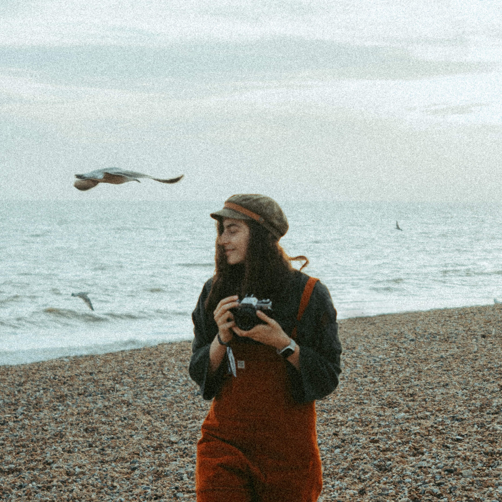 Analog photo of photographer Caro walking along the beach with her camera while pigeons fly past her