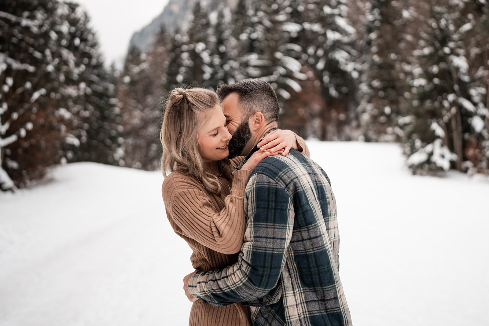 Engaged couple embracing in a snowy field while the man whispers into the woman’s ear