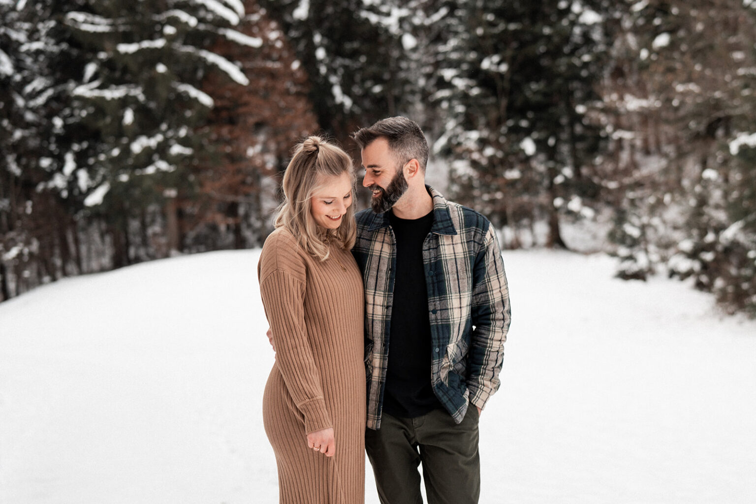 Couple standing in a snowy field while the man whispers into the woman’s ear