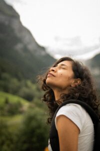 Young Indian woman standing in front of a stunning mountain landscape, facing the sky with closed eyes while enjoying a peaceful moment in nature