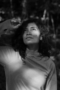 Vintage cinematic black-and-white photo of a young woman looking upward through her hands while light shines through the trees